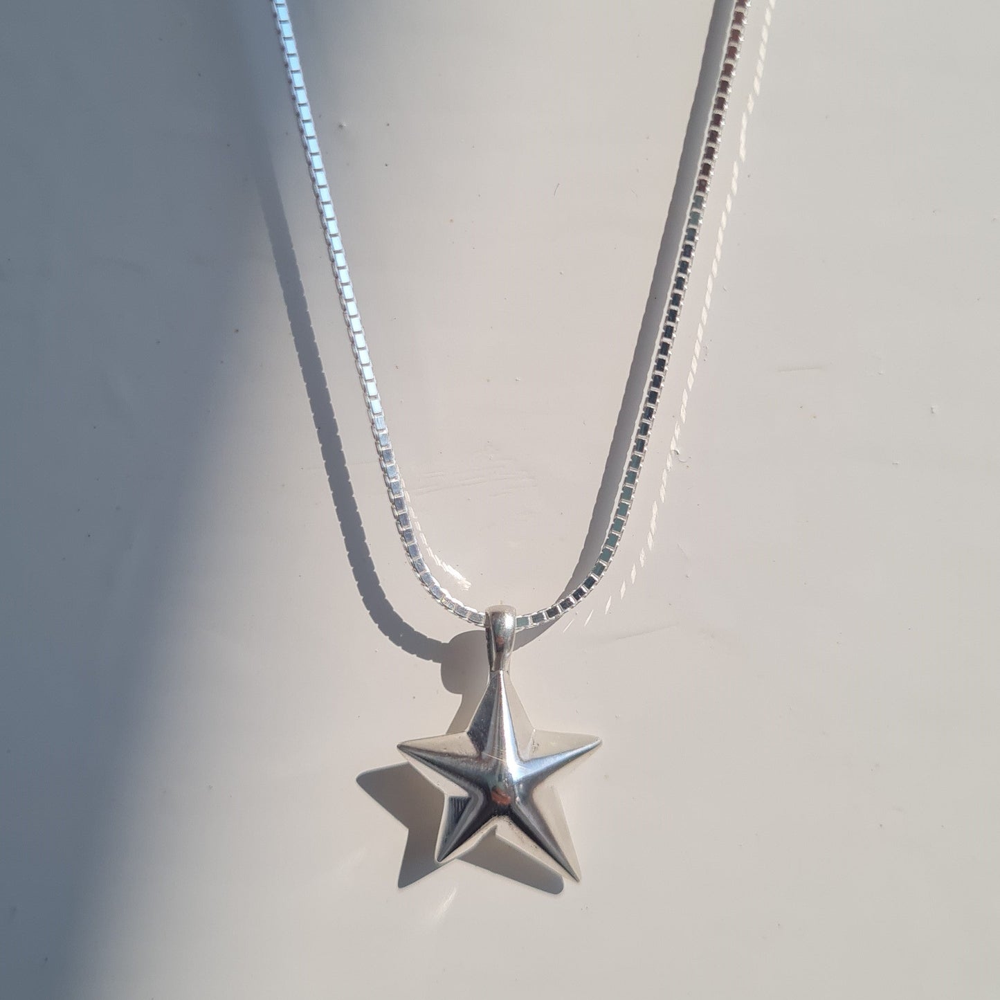 A silver star pendant necklace on a delicate silver chain, displayed against a light gray background with soft diagonal shadows.