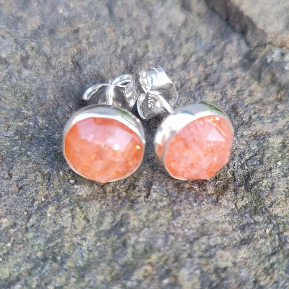 A pair of silver stud earrings with round orange-pink cabochon gemstones, displayed on a textured gray rock surface.