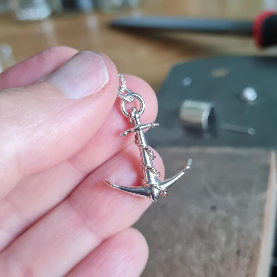 A small silver anchor charm with 9ct rose gold rope detail, held between a person's fingers, with a blurred workshop background