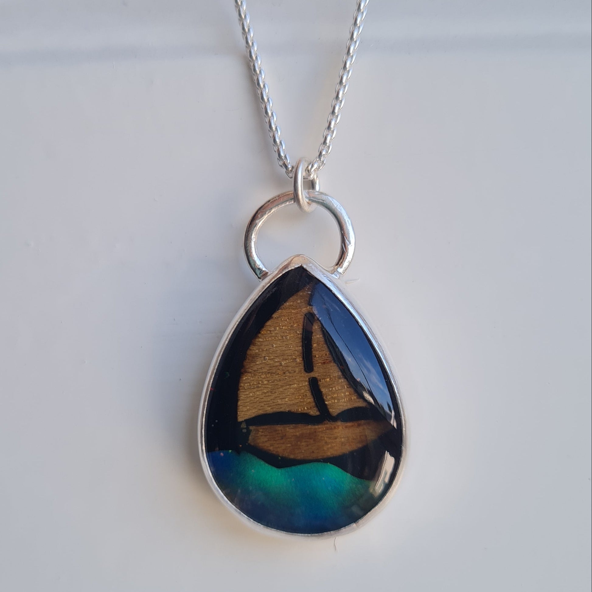 A silver teardrop pendant necklace featuring an iridescent aurora opal wave inlay with blue, green, and wood yacht inlay with black star sky background, suspended from a delicate silver chain