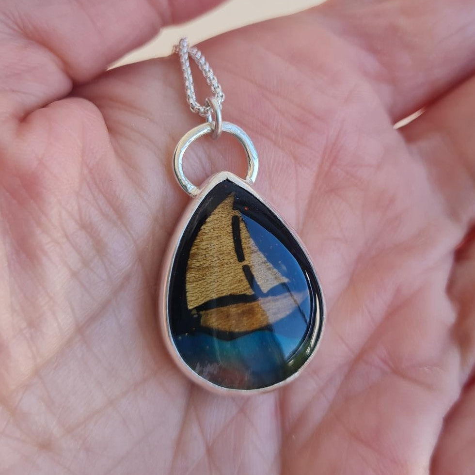 A silver teardrop pendant necklace featuring an aurora opal wave inlay, wood yacht inlay and black star sky, held in persons hand.