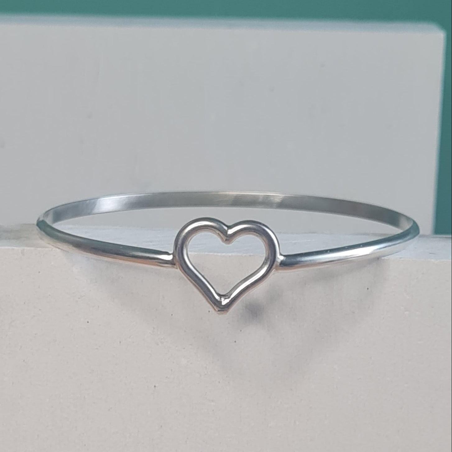 A solid silver bangle bracelet with a small open heart design in the center, shown resting against a white surface.