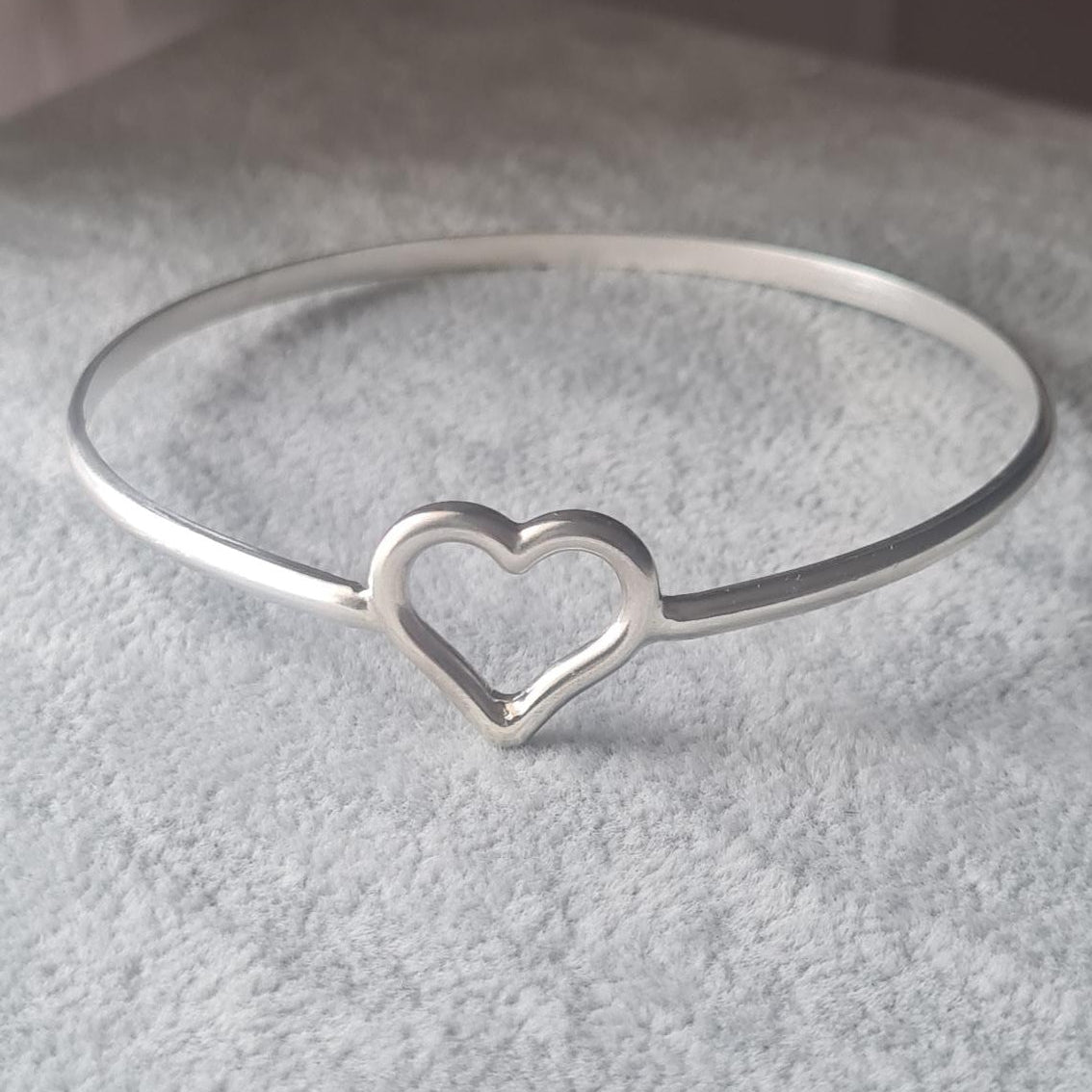 A silver bangle bracelet with a small open heart design, displayed on a textured gray stand.