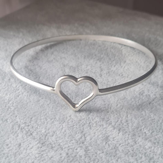 A silver bangle bracelet with a small open heart design, displayed on a textured gray stand.