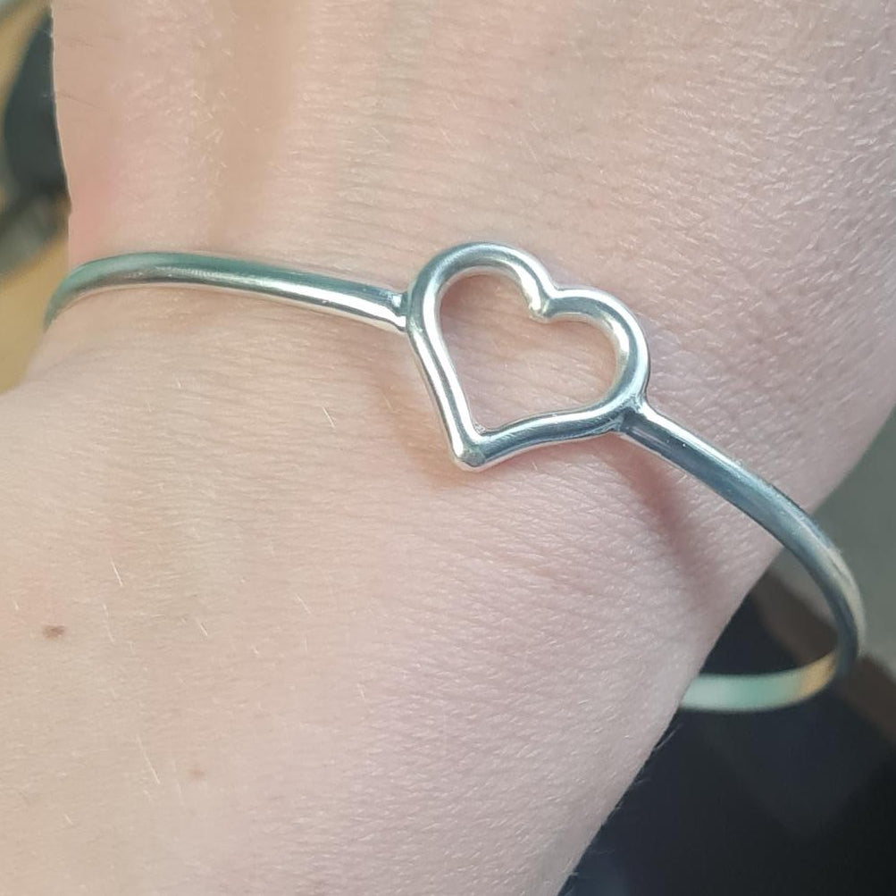 A solid silver bangle bracelet with an open heart design, shown worn on a person's wrist.