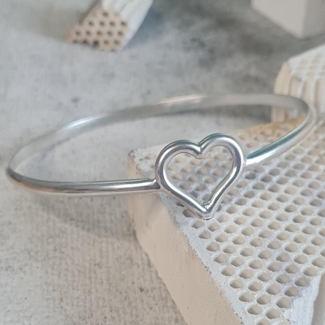 A solid silver bangle bracelet with a small heart-shaped cutout detail, displayed on a textured white surface.