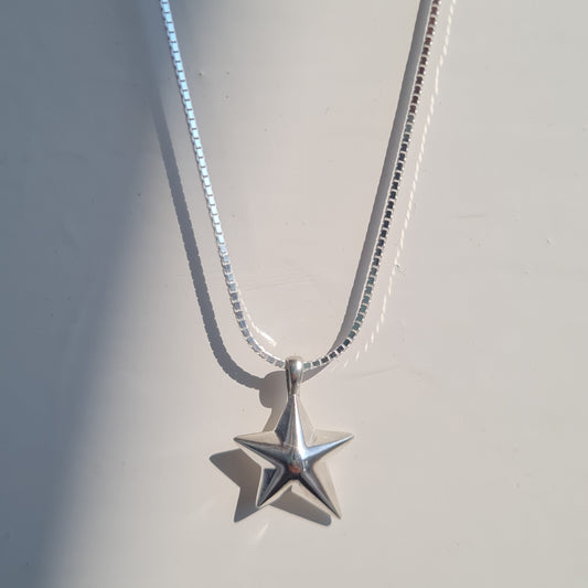 A silver star pendant necklace on a delicate silver chain, displayed against a light gray background with soft diagonal shadows.