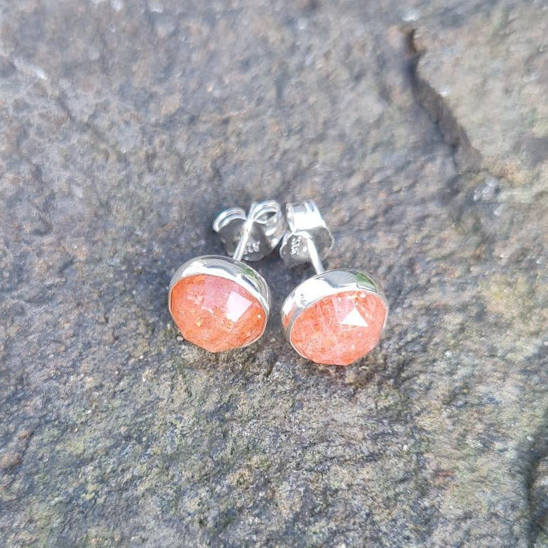 A pair of silver stud earrings with round orange-brown cabochon gemstones resting on a textured gray rock with green foliage blurred in the background.